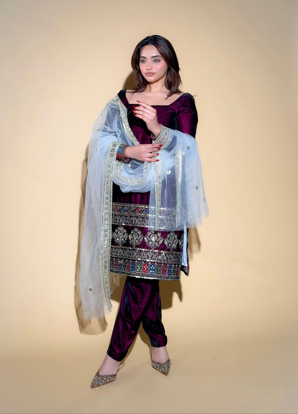 Woman wearing a traditional outfit with a purple top and pants, holding a matching dupatta against a beige background.