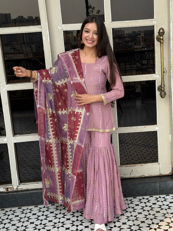 Woman wearing a long, patterned dress standing in front of a window with a decorative wall.
